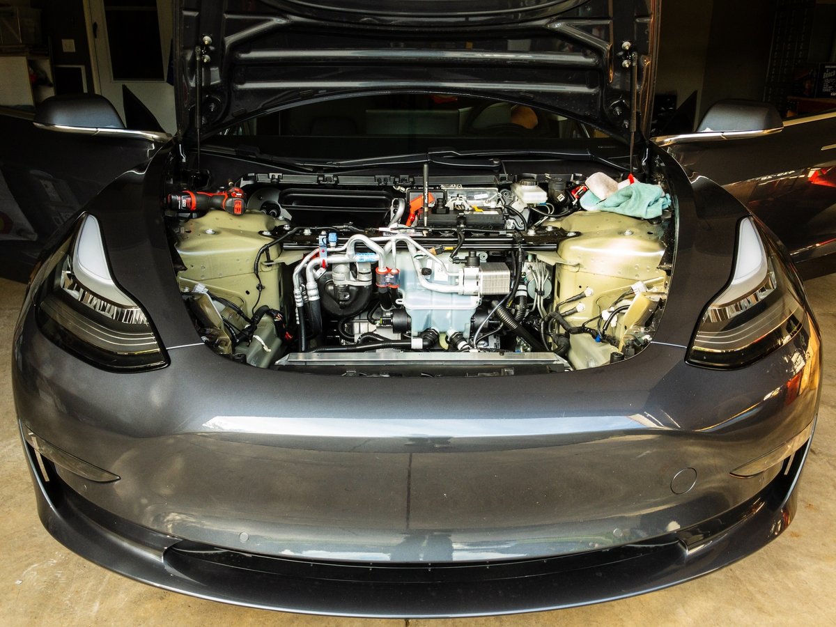A Tesla Model 3 has its front hood open and liner removed to reveal the firewall and various system components.