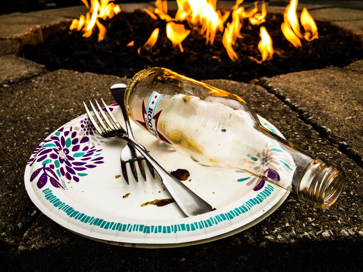 An empty root beer bottle and a post-meal paper plate rest on a stone wall of a firepit with a fire in the background